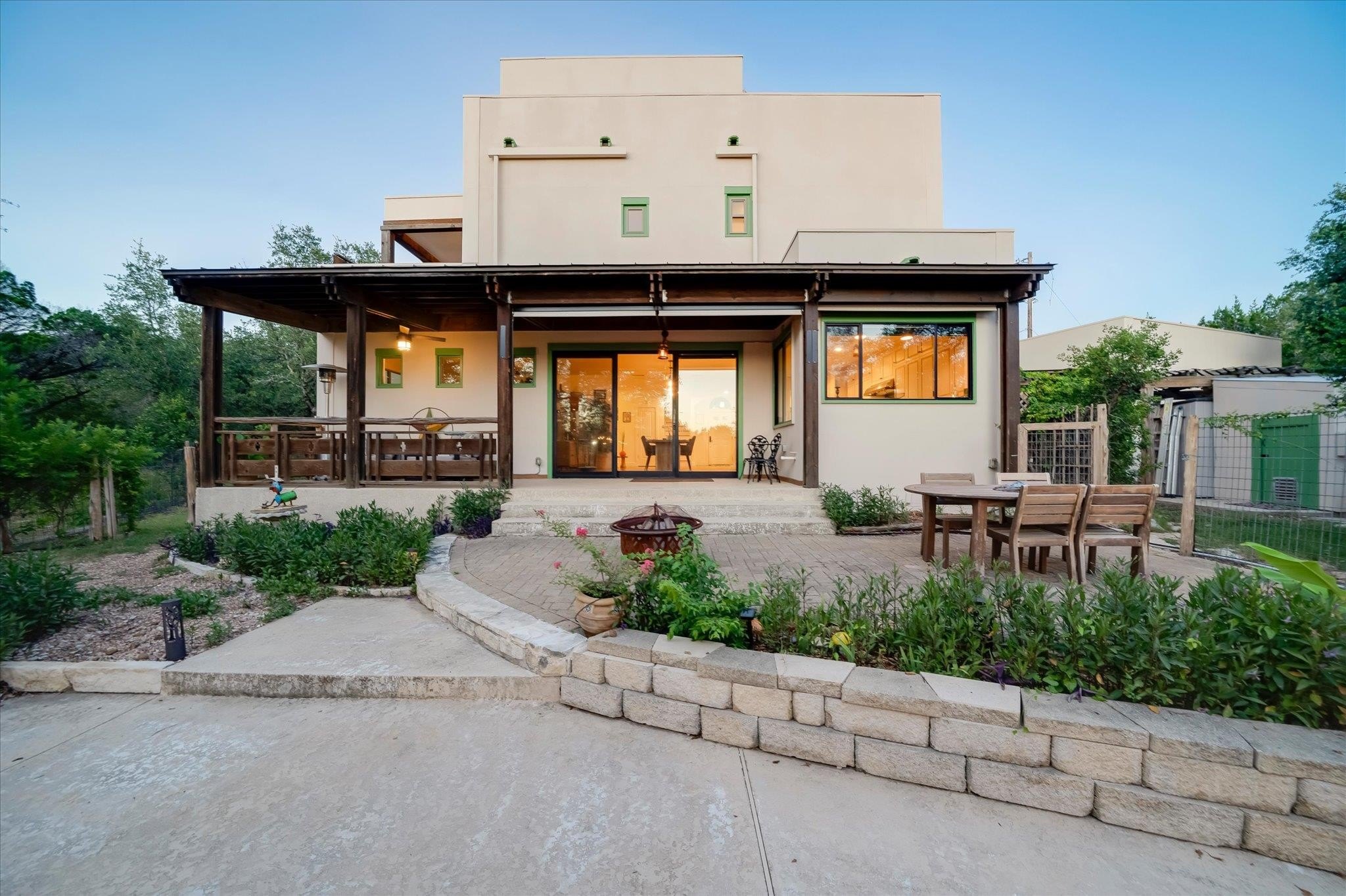 Cedar-columned back porch and stucco patio at 8300 Lime Creek Road, with a raised limestone garden bed and dining table at dusk
