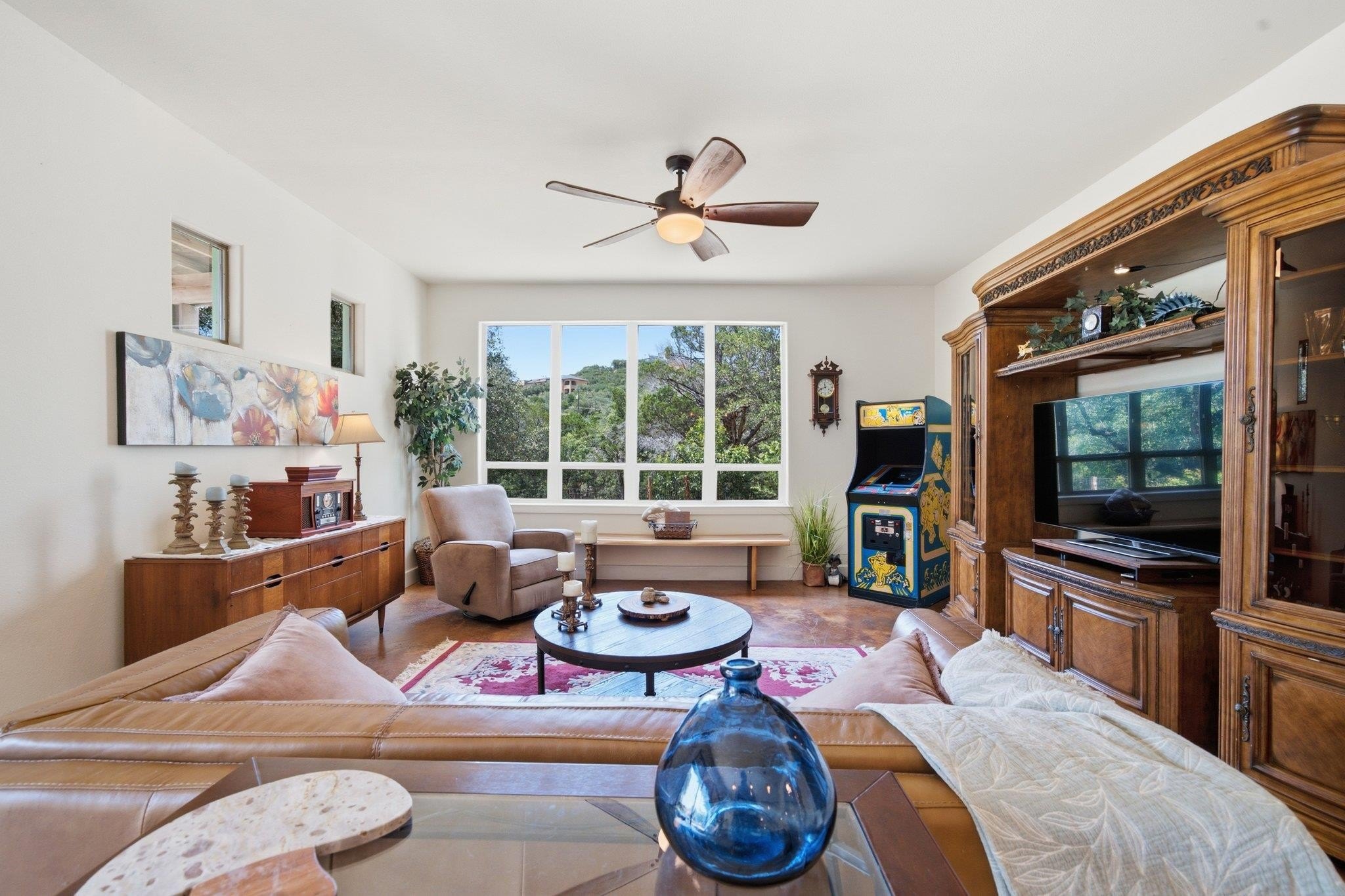Second sitting area at 8300 Lime Creek Road with a leather sofa, a large picture window framing the Hill Country woodland, and a vintage arcade cabinet against the far wall