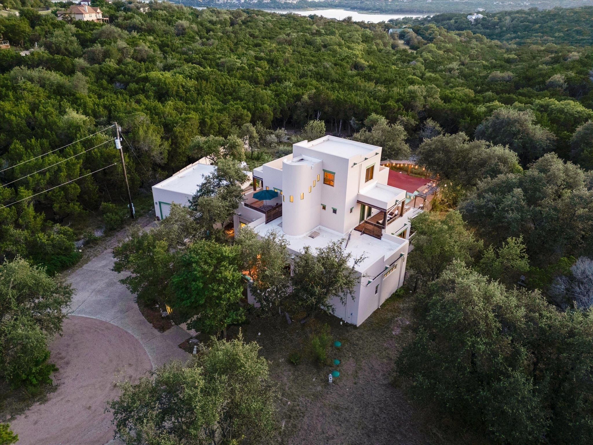Aerial dusk view of 8300 Lime Creek Road, a tiered white stucco home on a wooded ridge above Lake Travis in Volente, Texas